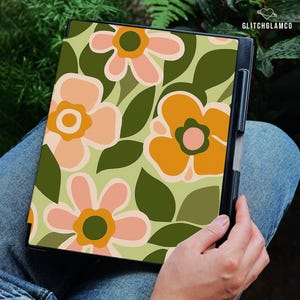 May include: A black tablet case featuring a retro floral pattern in peach, orange, and green on a light green background. The case is held by a person wearing blue jeans. The text "GLITCHGLAMCO" is visible in the upper right corner.
