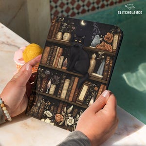 Puede incluir: Una funda para tableta con un gato negro sobre un diseño de estantería. La funda presenta un fondo oscuro con libros, flores y una luna creciente. El texto "GLITCHGLAMCO" está en la esquina superior derecha.