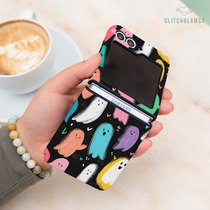 May include: Black phone case featuring a pattern of colourful ghosts. The case is open, displaying the phone screen and the Samsung logo. The case has a pink, yellow, and green border. The background is black with ghosts in various colours.