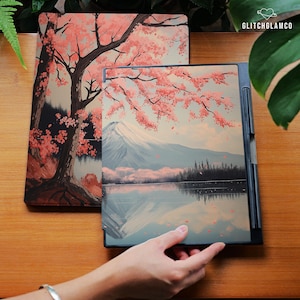 以下が含まれることがあります： 日本の風景画が描かれた2つのデジタルタブレットカバー。1つは桜の木、もう1つは湖のある山岳風景です。カバーはマット仕上げで、木製の表面に展示されています。