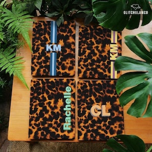 May include: Four rectangular covers featuring a brown and black leopard print pattern. Each cover displays a vertical stripe with personalized text: KM in light blue, KM in yellow, Rachelle in light green, and CL in light orange.