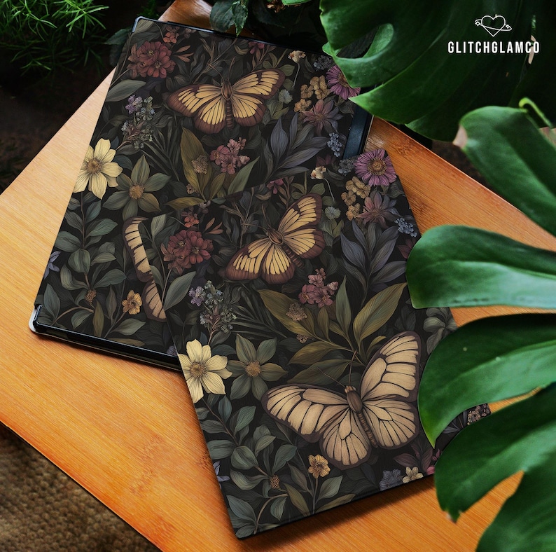 May include: A dark-colored tablet case featuring a botanical design with butterflies and flowers in shades of yellow, brown, and purple. The case is set on a wooden surface, with the GlitchGlamCo logo visible.