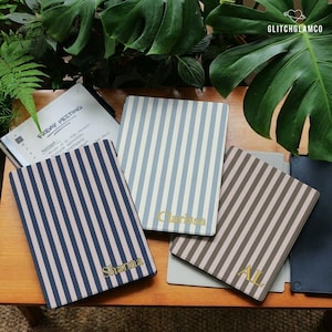 Può includere: Quaderni a righe verticali blu navy e beige, azzurro e bianco, e marrone e beige. Ogni quaderno ha un nome o delle iniziali color oro. Un quaderno è aperto, mostrando il testo "FRIDAY MEETING". Lo sfondo include piante verdi.