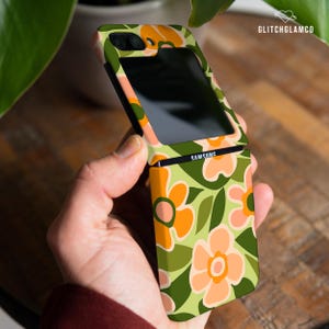 May include: A green, orange, and pink floral phone case for a Samsung Galaxy Z Flip phone. The case has a retro 70s style.