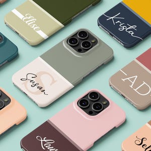 May include: A collection of personalized phone cases in various colors, including teal, pink, and olive green. Each case features a two-tone design with a name or initials in a stylish font. The cases are arranged on a light blue surface.