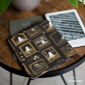 May include: A black Kindle cover featuring a repeating pattern of framed ghost illustrations. The frames are gold, and the ghosts are white. The cover is on a wooden table next to a Kindle device.