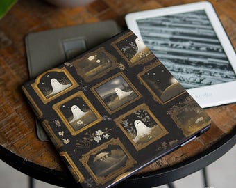 Peut inclure: Une housse de Kindle noire avec un motif répété d'illustrations de fantômes encadrées. Les cadres sont dorés et les fantômes blancs. La housse est sur une table en bois à côté d'un appareil Kindle.