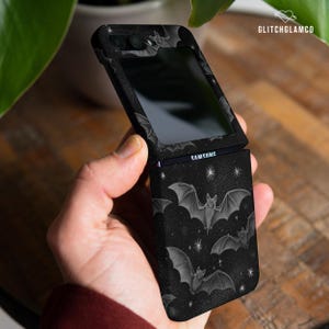 Puede incluir: Funda negra para teléfono Samsung con un diseño de murciélagos y estrellas. La funda está abierta, mostrando la pantalla del teléfono. La funda tiene un fondo oscuro con un patrón repetido de murciélagos y estrellas.