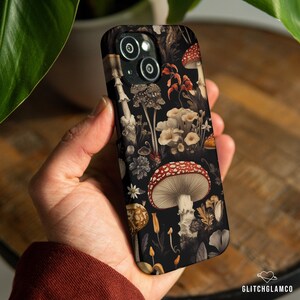 May include: A black phone case featuring a detailed mushroom pattern in shades of red, white, and beige against a dark background. The design includes various mushroom species and floral elements, creating a botanical theme. The case is held in a hand.
