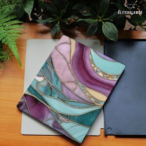 May include: A decorative tablet case with a stained glass design. The case features a vibrant color palette of teal, purple, pink, and gold, with a flowing, abstract pattern. The GlitchGlamCo logo is visible in the top right corner.