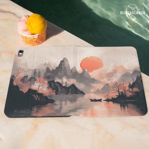 May include: An iPad case featuring a serene landscape painting with mountains, a lake, and a setting sun in shades of gray, peach, and white. A small boat is on the lake. The case is on a marble surface next to a cup of ice cream.