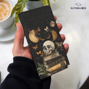 Puede incluir: Una funda de teléfono de cuero negro con un diseño de calavera y mariposa. La calavera está sentada sobre una pila de libros antiguos con detalles dorados. El fondo es un cielo estrellado oscuro con una luna creciente.