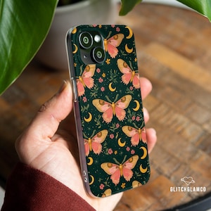 Può includere: Una custodia per telefono con sfondo verde scuro con un motivo ripetuto di farfalle rosa e gialle, lune crescenti e piccoli fiori rosa. La custodia ha un bordo trasparente.