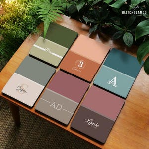 Może przedstawiać: Zestaw spersonalizowanych podkładek w różnych kolorach, w tym zielonym, brzoskwiniowym, turkusowym i bordowym. Każda podkładka zawiera imię lub inicjał w stylowej czcionce. Podkładki ułożone są na drewnianej powierzchni, z zielenią w tle.