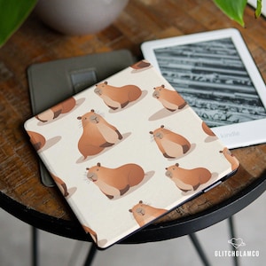 May include: A Kindle case featuring a repeating pattern of cartoon capybaras in shades of brown and tan on a cream background. The case is open, revealing the screen of a Kindle e-reader. A gray case and a Kindle are also visible.
