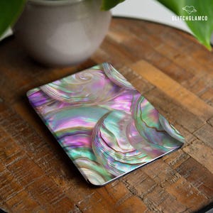 May include: A rectangular decorative item with an iridescent, swirling pattern in shades of purple, green, and pink. The design resembles the inside of a seashell. The item is placed on a wooden surface.