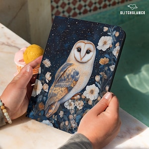 May include: A notebook with a dark blue cover featuring a detailed illustration of a barn owl surrounded by white and yellow flowers. The owl has a white face and chest with brown and blue wing details. The text "GLITCHGLAMCO" is in the upper right corner.