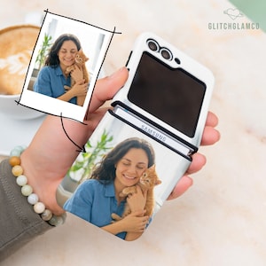 May include: White Samsung flip phone case with a photo of a woman holding a ginger kitten. The case is open, revealing the screen and the Samsung logo. A photo of the same image is in the upper left corner. The phone is held in a hand.