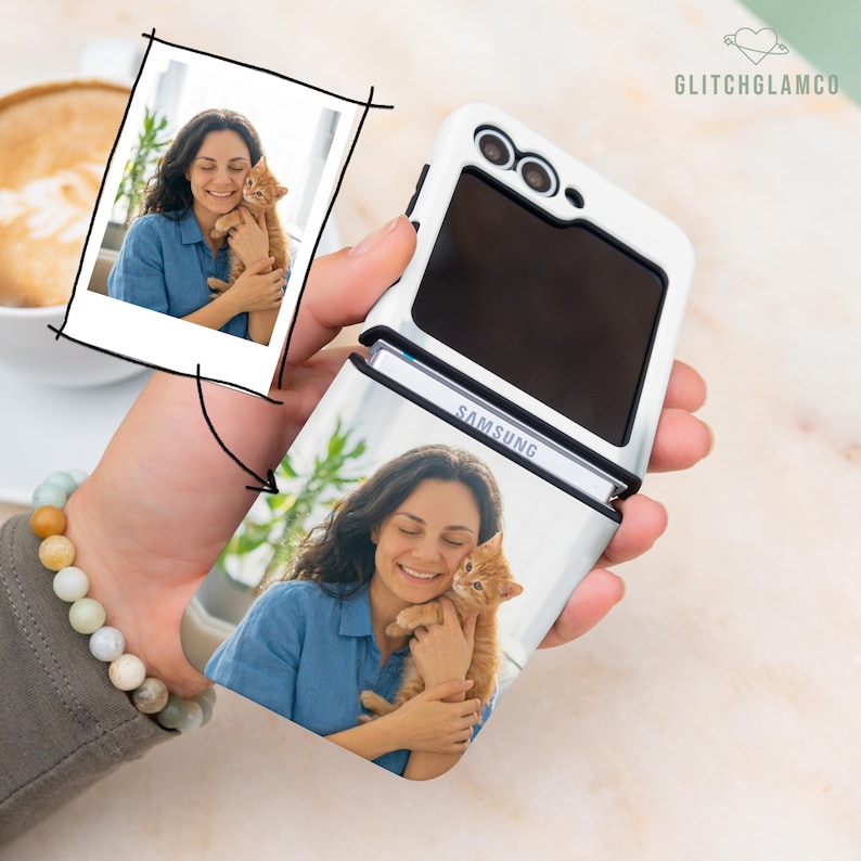 May include: White Samsung flip phone case with a photo of a woman holding a ginger kitten. The case is open, revealing the screen and the Samsung logo. A photo of the same image is in the upper left corner. The phone is held in a hand.