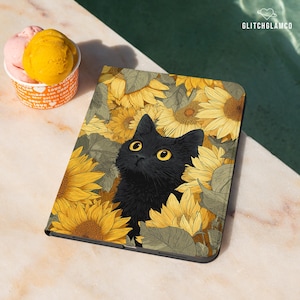 May include: A black cat with yellow eyes peeks out from a field of yellow sunflowers on a tablet case. The case has a black border. A cup of gelato with pink and yellow scoops sits nearby.