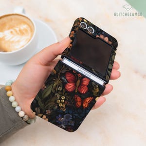 May include: A black Samsung phone case featuring a floral and butterfly design. The case displays red and orange butterflies amidst a pattern of flowers and foliage. The phone is partially open, showing the screen and the Samsung logo.