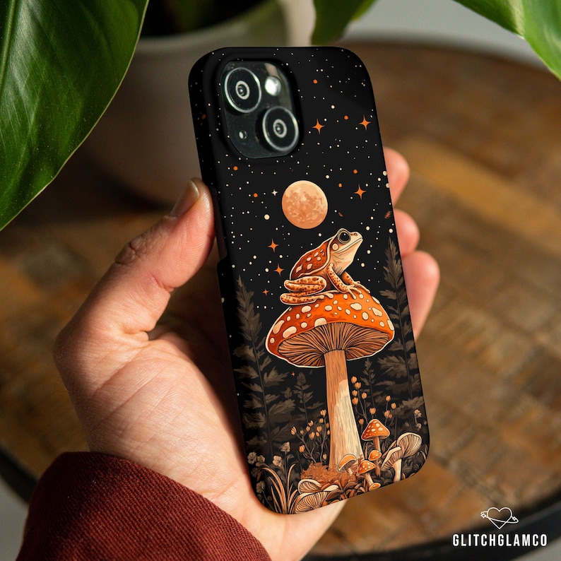 May include: Black phone case featuring a whimsical illustration of a frog atop a mushroom against a starry night sky. The design includes a full moon, stars, and forest elements. The case has a glossy finish and is held in a hand.