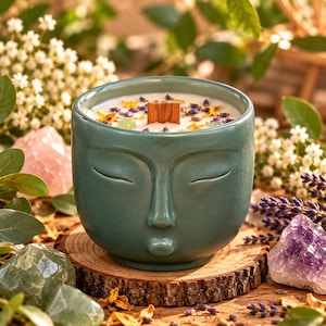 May include: A teal ceramic candle vessel shaped like a human face with closed eyes and pursed lips. The candle features white wax, a wooden wick, and is topped with dried lavender buds, yellow flower petals, and small green crystal stones.
