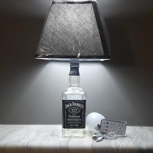 Może przedstawiać: Unikalna lampa wykonana z butelki whisky Jack Daniel's, zwieńczona czarnym abażurem. Zawiera białą żarówkę, pilota i kabel USB. Lampa stoi na białej marmurowej powierzchni.