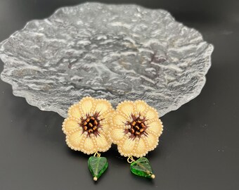 May include: A pair of delicate earrings featuring embroidered cream-colored flowers with brown centers and green glass leaves dangling below.