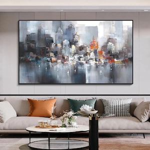 May include: Abstract cityscape painting in a black frame. The artwork depicts a skyline with buildings in shades of grey, blue, and orange, reflected in the water below. Small white sailboats dot the water.