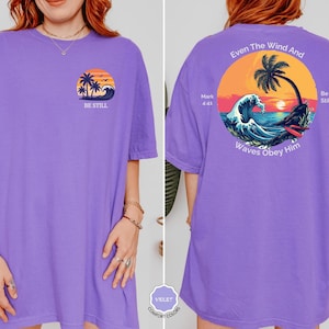 May include: A violet-colored t-shirt with a front graphic of a sunset, palm trees, and waves, with the text "BE STILL". The back features a circular design with a sunset, palm tree, waves, and the text "Even The Wind And Waves Obey Him".