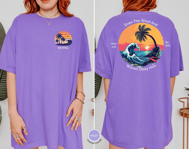 May include: A violet-colored t-shirt with a front graphic of a sunset, palm trees, and waves, with the text "BE STILL". The back features a circular design with a sunset, palm tree, waves, and the text "Even The Wind And Waves Obey Him".