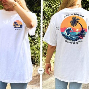 May include: White t-shirt with a beach-themed graphic. The front features a sunset, palm trees, and the words "BE STILL." The back displays a circular design with a palm tree, waves, and the text "Even The Wind And Waves Obey Him."