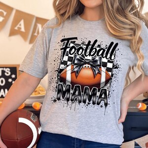 An athletic heather Football Mama graphic t shirt featuring a stylish football design with a checkered pattern and a bow.