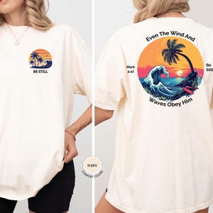 May include: Ivory-colored t-shirt with a beach-themed graphic on the front and back. The front features a sunset, palm trees, and the words "BE STILL." The back displays a circular design with waves, a palm tree, and the text "Even The Wind And Waves Obey Him."