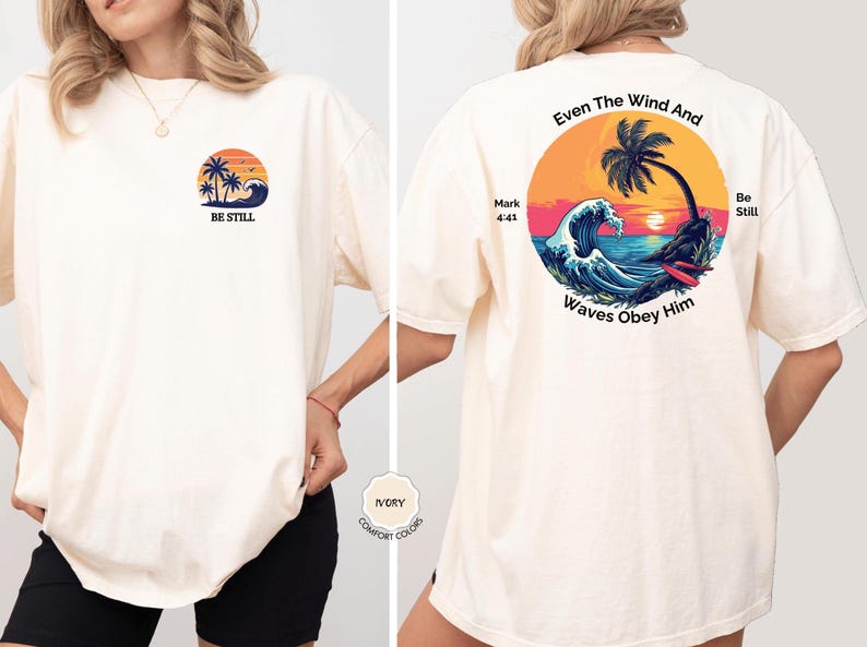 May include: Ivory-colored t-shirt with a beach-themed graphic on the front and back. The front features a sunset, palm trees, and the words "BE STILL." The back displays a circular design with waves, a palm tree, and the text "Even The Wind And Waves Obey Him."