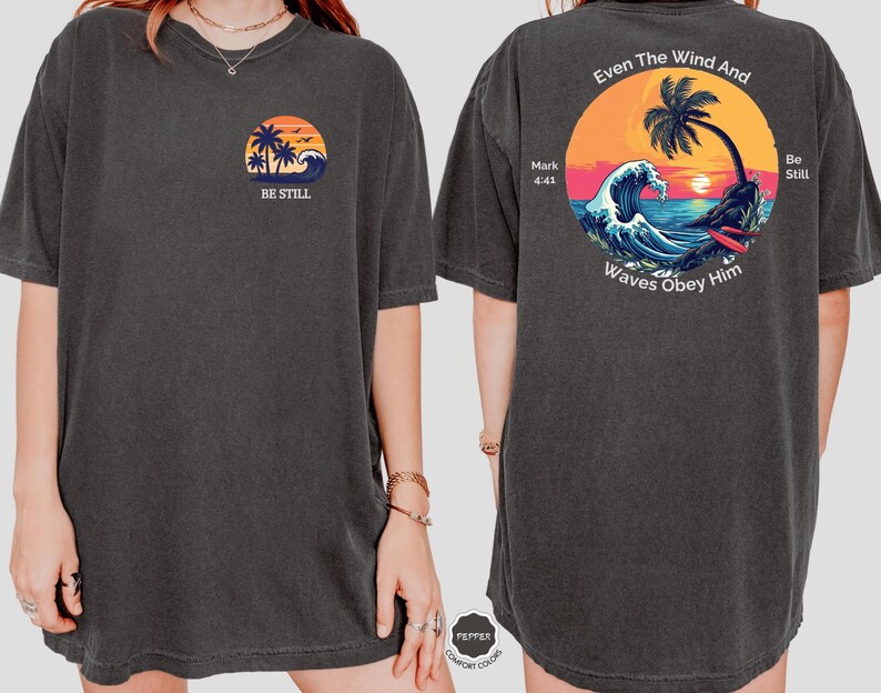 May include: Dark gray t-shirt with a front graphic of a sunset, palm trees, and the words "BE STILL." The back features a circular design with a palm tree, waves, and the text "Even The Wind And Waves Obey Him."