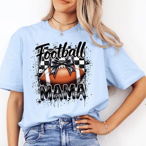 A light blue Football Mama graphic t shirt featuring a stylish football design with a checkered pattern and a bow.