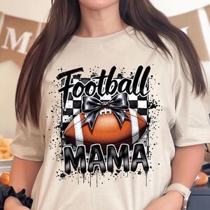 A sand Football Mama graphic t shirt featuring a stylish football design with a checkered pattern and a bow.