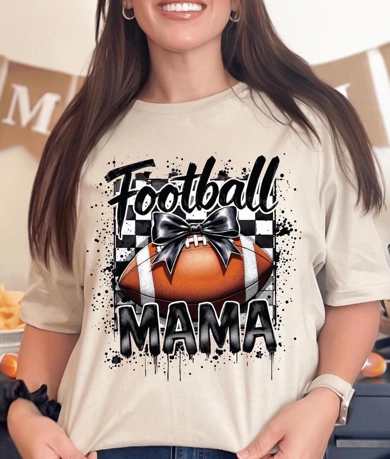 A sand Football Mama graphic t shirt featuring a stylish football design with a checkered pattern and a bow.