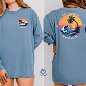 May include: A long-sleeved blue shirt with a front graphic of a sunset, palm trees, and waves, with the text "BE STILL." The back of the shirt features a larger graphic with the text "Even The Wind And Waves Obey Him."