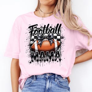 A light pink Football Mama graphic t shirt featuring a stylish football design with a checkered pattern and a bow.