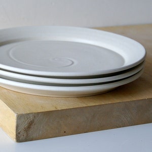 May include: Three white ceramic plates with a spiral design are stacked on a wooden surface. A small deer figurine stands nearby. The plates have a simple, minimalist design, suitable for a modern kitchen.
