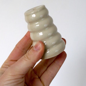 Peut inclure: Un salière en céramique de couleur crème avec un motif en spirale. Le salière est tenu dans une main sur un fond blanc. Le salière a un petit trou en haut pour distribuer le sel.