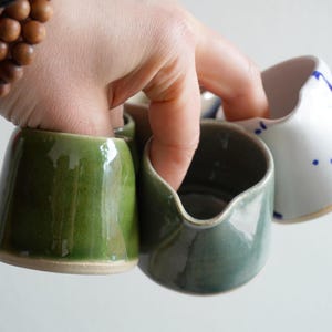 Peut inclure: Une collection de petits pichets en céramique avec des finitions lisses et brillantes dans des tons vert olive, vert sauge et blanc avec des taches bleues. Chaque pichet présente un corps arrondi, un petit bec verseur et une base non émaillée.