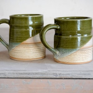 May include: Two ceramic mugs featuring a two-tone design with a glossy olive green glaze on the upper portion and a textured, unglazed beige finish on the lower section. Each mug has a curved handle and a cylindrical body with a distinct horizontal ridge.