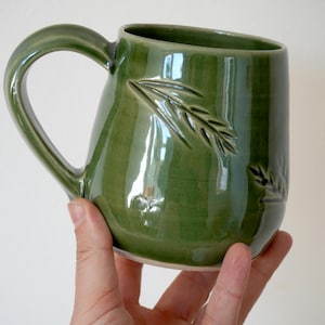 May include: A glossy olive green ceramic mug featuring a curved handle and a debossed wheat stalk pattern on the exterior surface. The mug has a smooth, reflective finish and a rounded body shape, showcasing a consistent deep green glaze throughout.