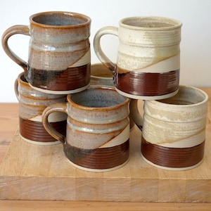 May include: Six handmade ceramic mugs in a stack. Each mug has a unique design with brown, beige, and grey tones. The mugs have handles and are arranged on a wooden surface.