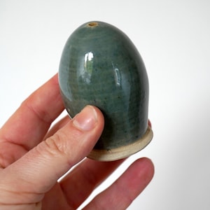 Peut inclure: Un salière en céramique en forme d'œuf, de couleur vert-bleu brillant. La base est beige clair et le dessus est percé d'un petit trou. La pièce en céramique est lisse et arrondie, et est tenue dans une main.