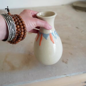 May include: A small, cream-coloured ceramic vase with a narrow neck and a rounded body. The upper portion of the vase features decorative drip patterns in muted blue and bright orange glaze, creating a contrast against the smooth, light surface.